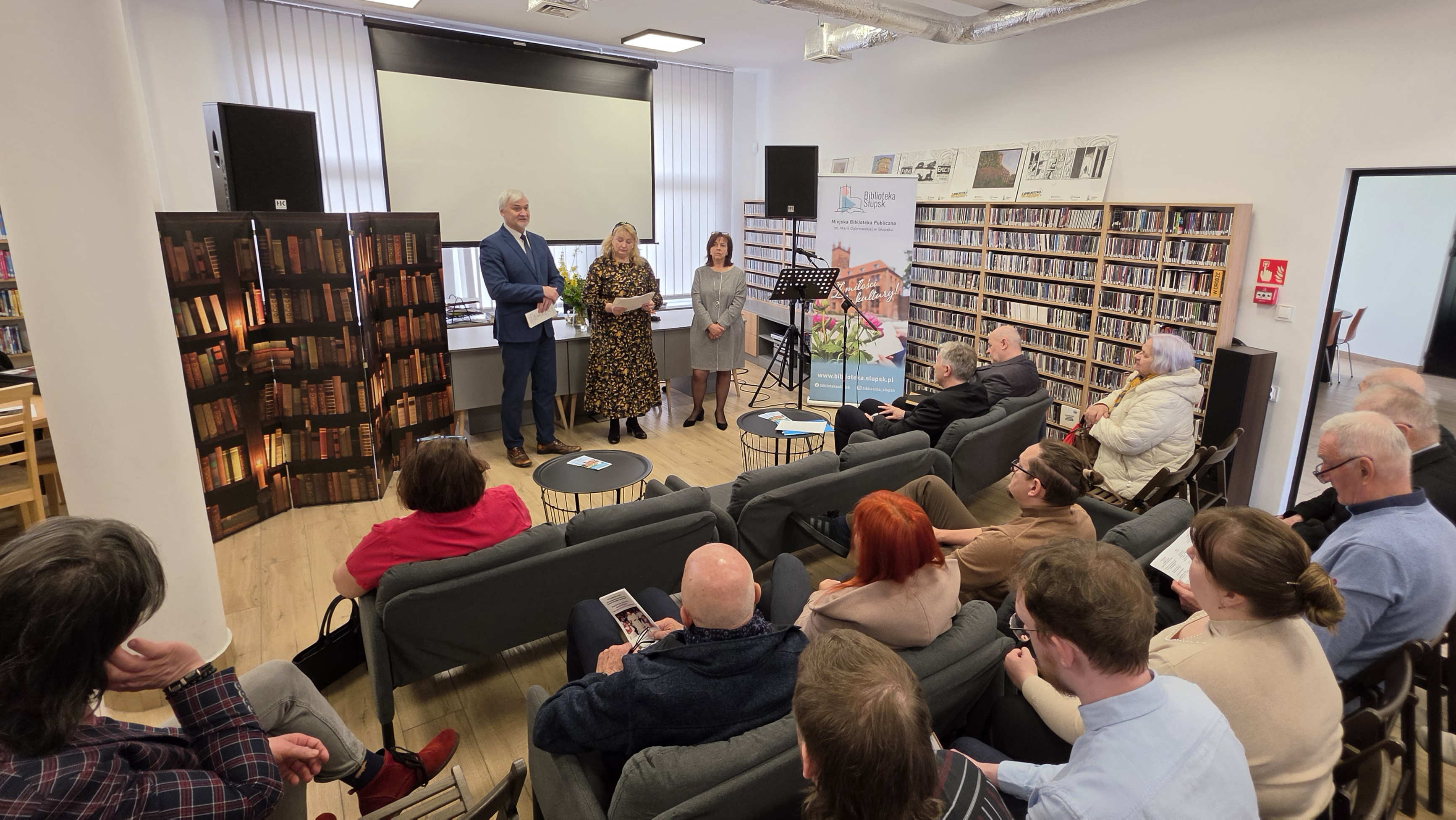 zdjęcie z góry, na pierwszym planie siedząca publiczność na kanapach, w oddali 3 osoby stojące