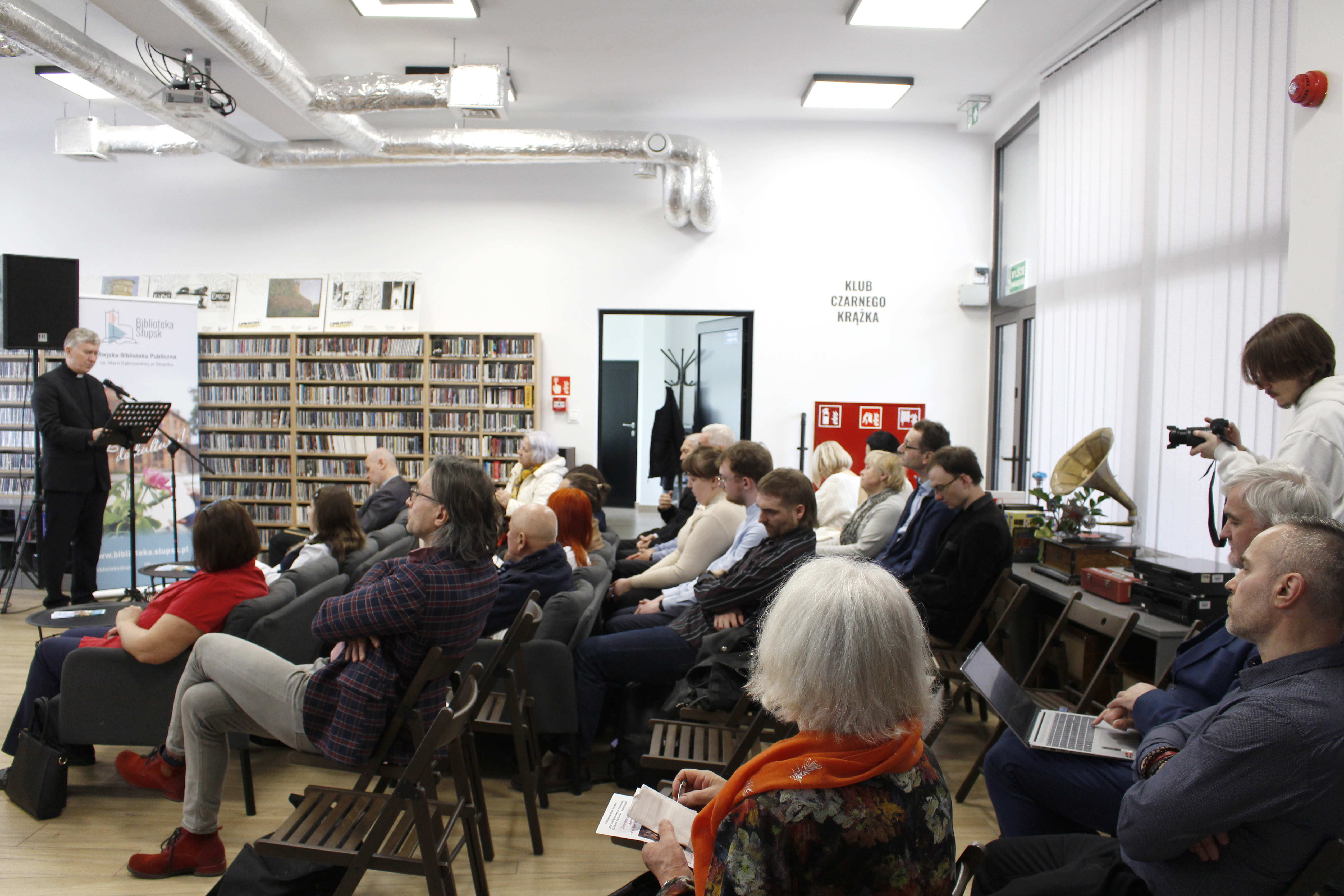 publiczność siedząca i słuchająca wypowiedzi stojącego po lewej stronie przy mikrofonie Księdza