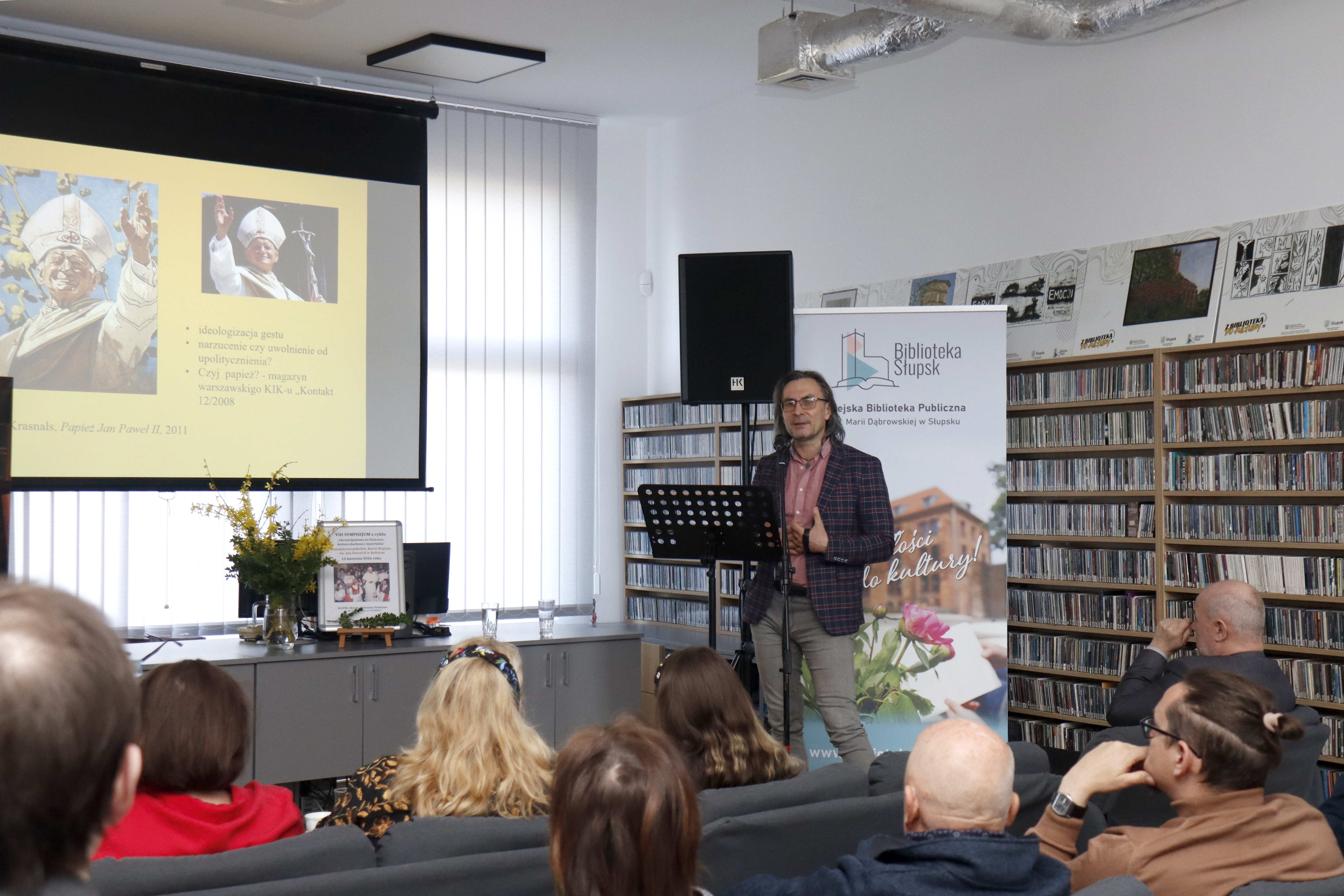 na pierwszym planie głowy publiczności siedzącej na kanapach, zwróconych w stronę stojącego na dalszym planie mężczyzny przemawiającego przez mikrofon, w tle po lewej stronie widnieje prezentacja ze zdjęciami Papieża Jana Pawła II