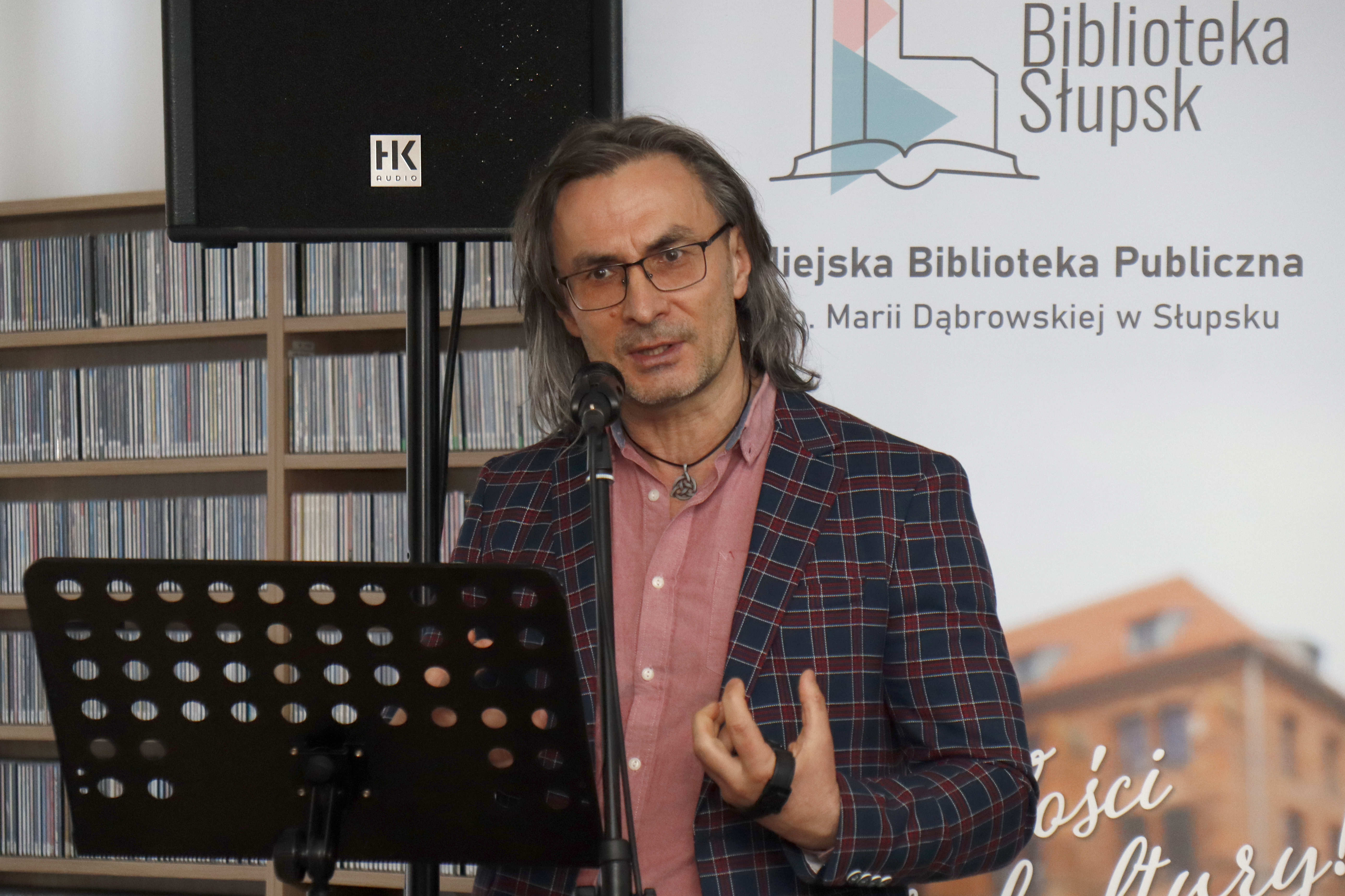 zbliżenie mężczyzny przemawiającego przez mikrofon, na tle regałów z płytami i rollupu bibliotecznego
