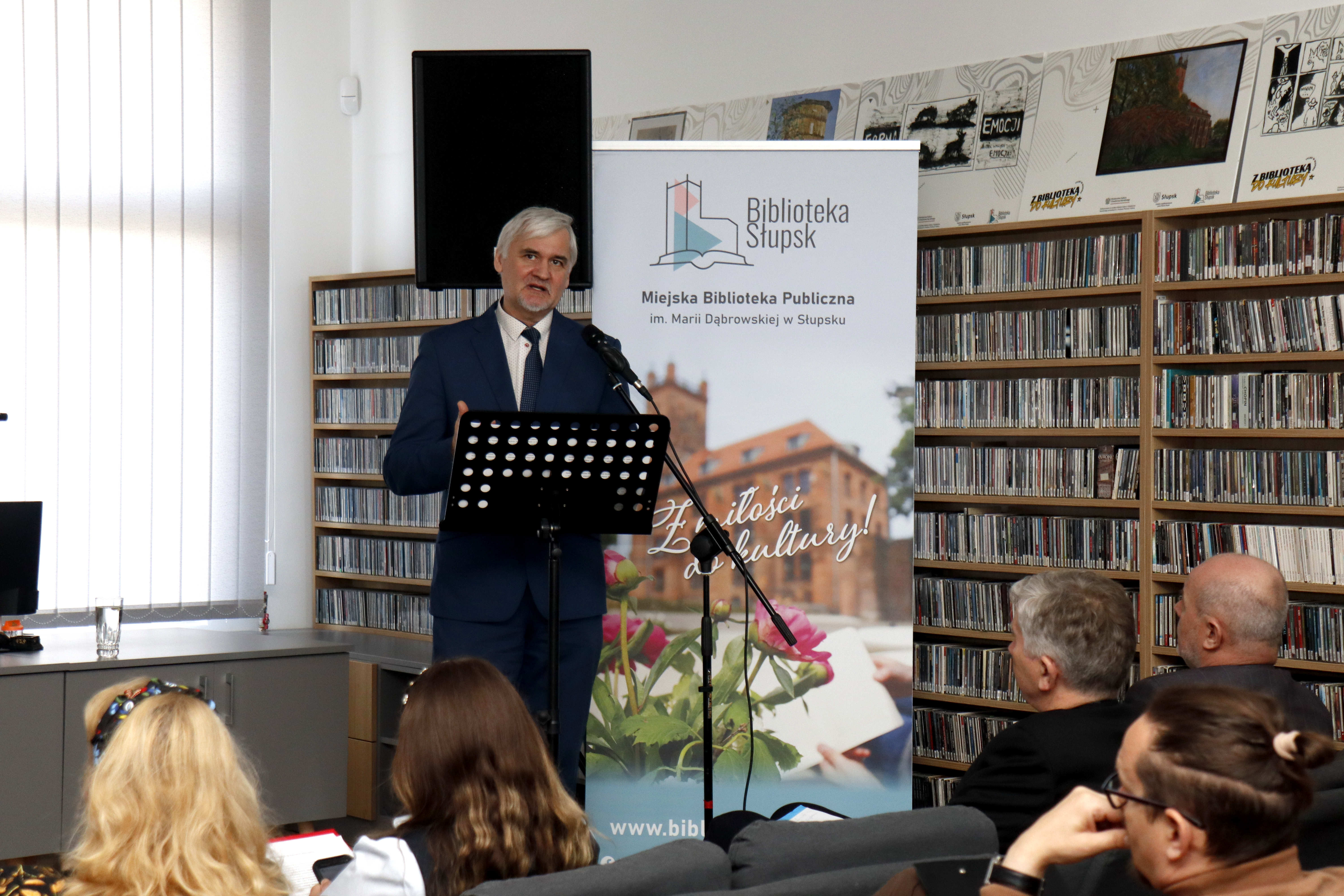 Sympozjum, mężczyzna stoi przy mikrofonie, publiczność tyłem, słucha