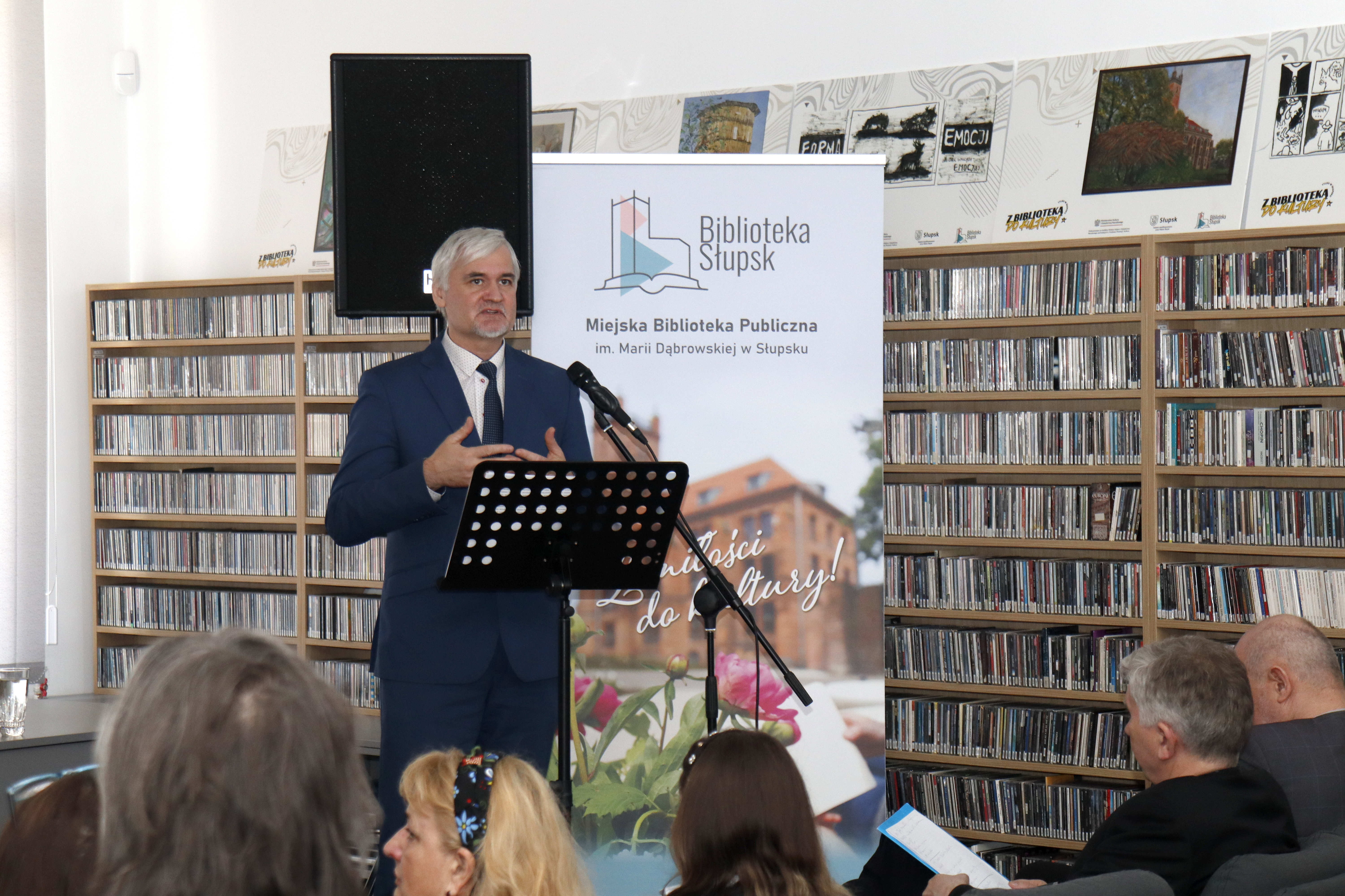 mężczyzna trzymający ręce na wysokości piersi, przy mikrofonie i na tle regałów z płytami i rollupie bibliotecznym