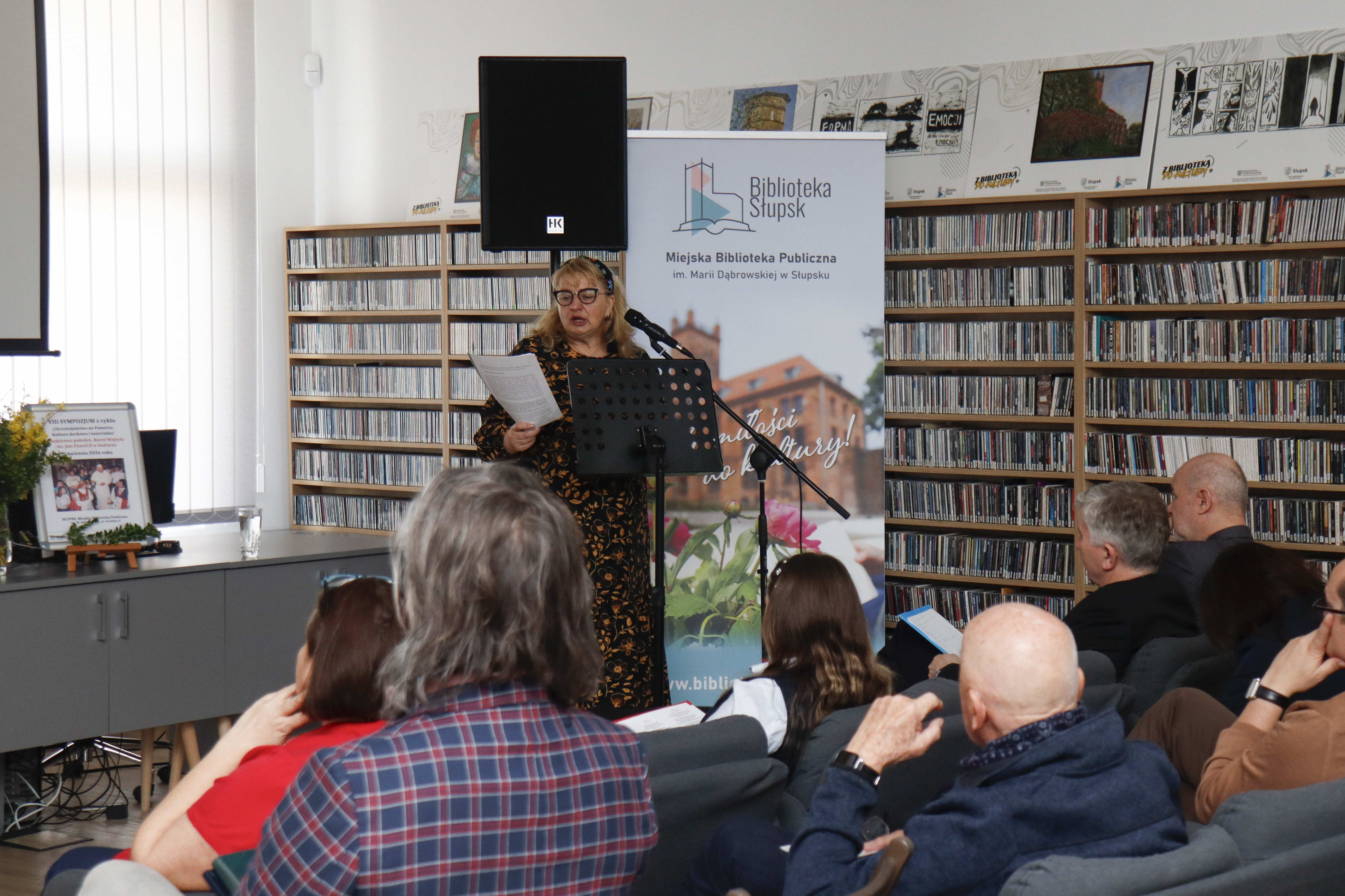 na pierwszym planie głowy publicznośći zwróconych w stronę przemawiającej kobiety stojącej przy mikrofonie i trzymającej w ręce papiery, czyta z nich