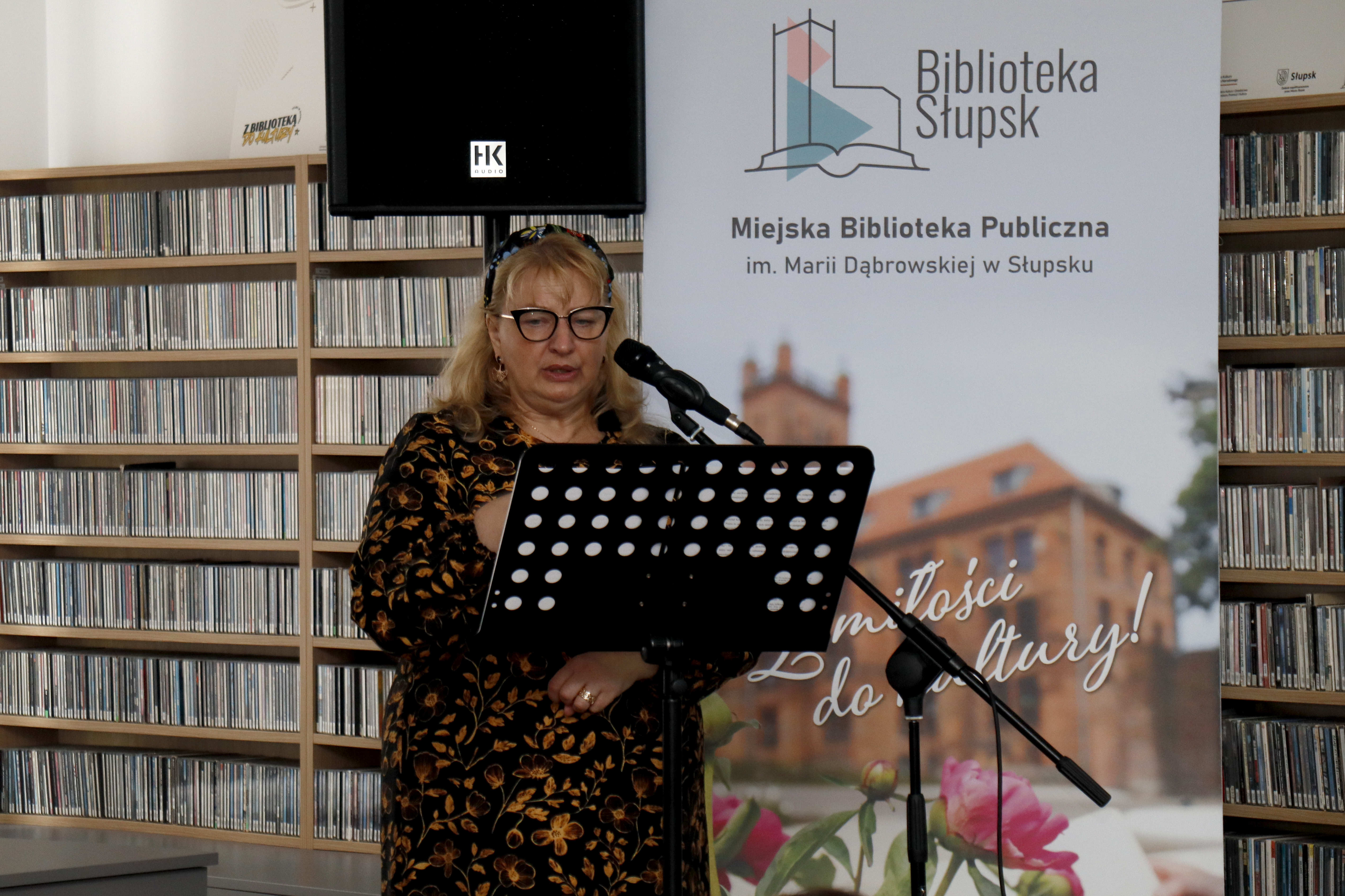 Kobieta stojąca przy mikrofonie, w tle rollup biblioteczny i regały z płytami