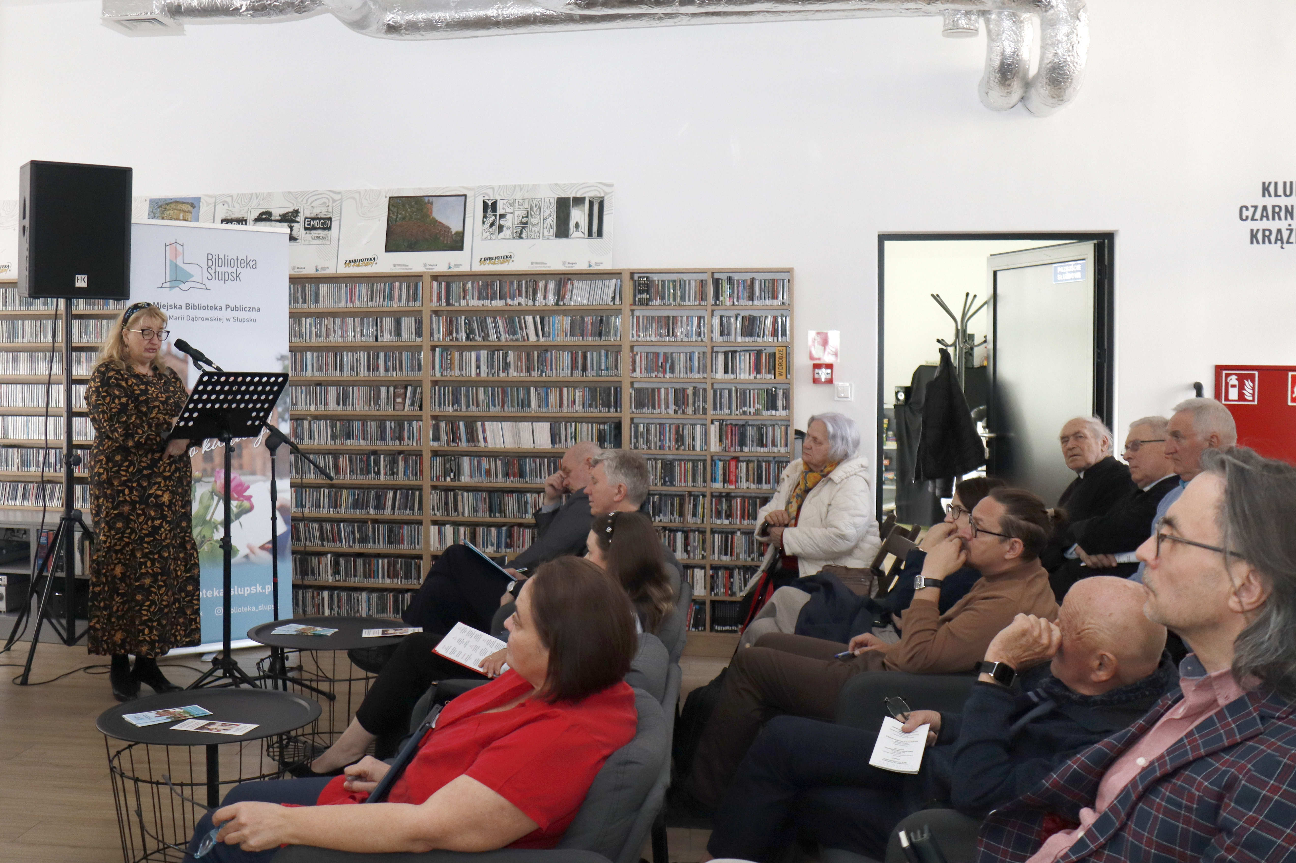 publiczność siedząca na kanapach, sfotografowana z boku, po lewej kobieta przemawiająca do mikrofonu