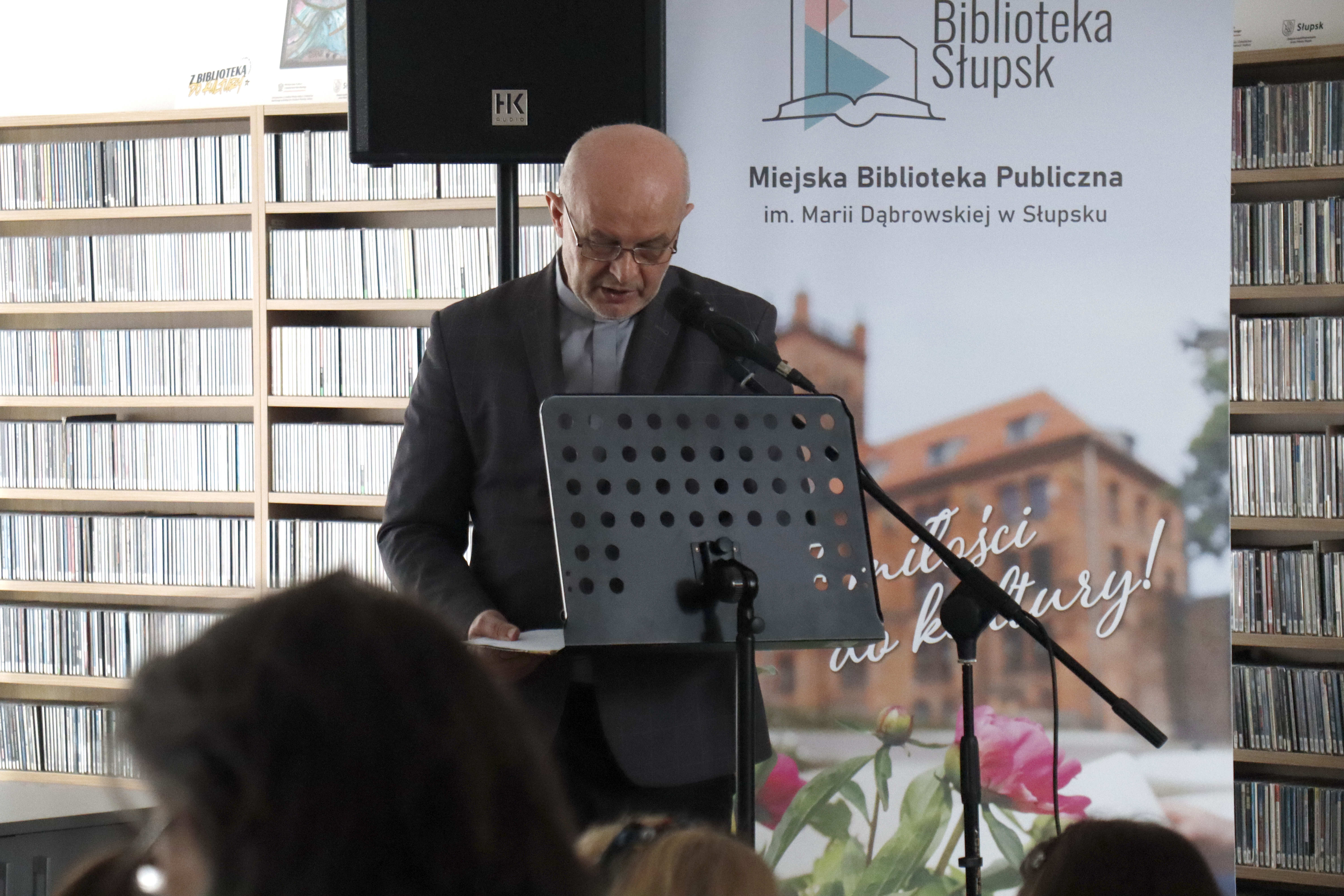 ksiądz stojący przy mikrofonie na tle regałów z płytami i roll-upu bibliotecznego