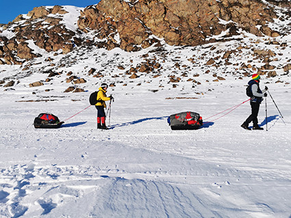 para osób ciągnących po śniegu na sankach bagaże, w tle ośnieżone góry
